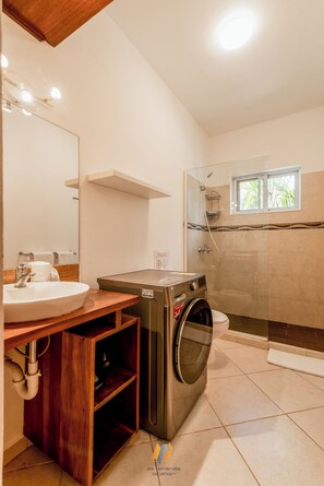 Baño con lavamanos, espejo, tocador de madera, lavadora, ducha y ventana con vista a palmeras.