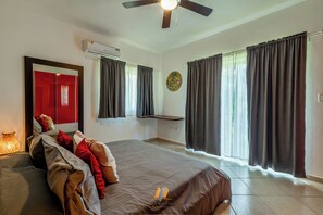 Habitación de hotel con cama, ventilador de techo, equipo de aire acondicionado, ventana con cortinas y una obra de arte colgada en la pared.