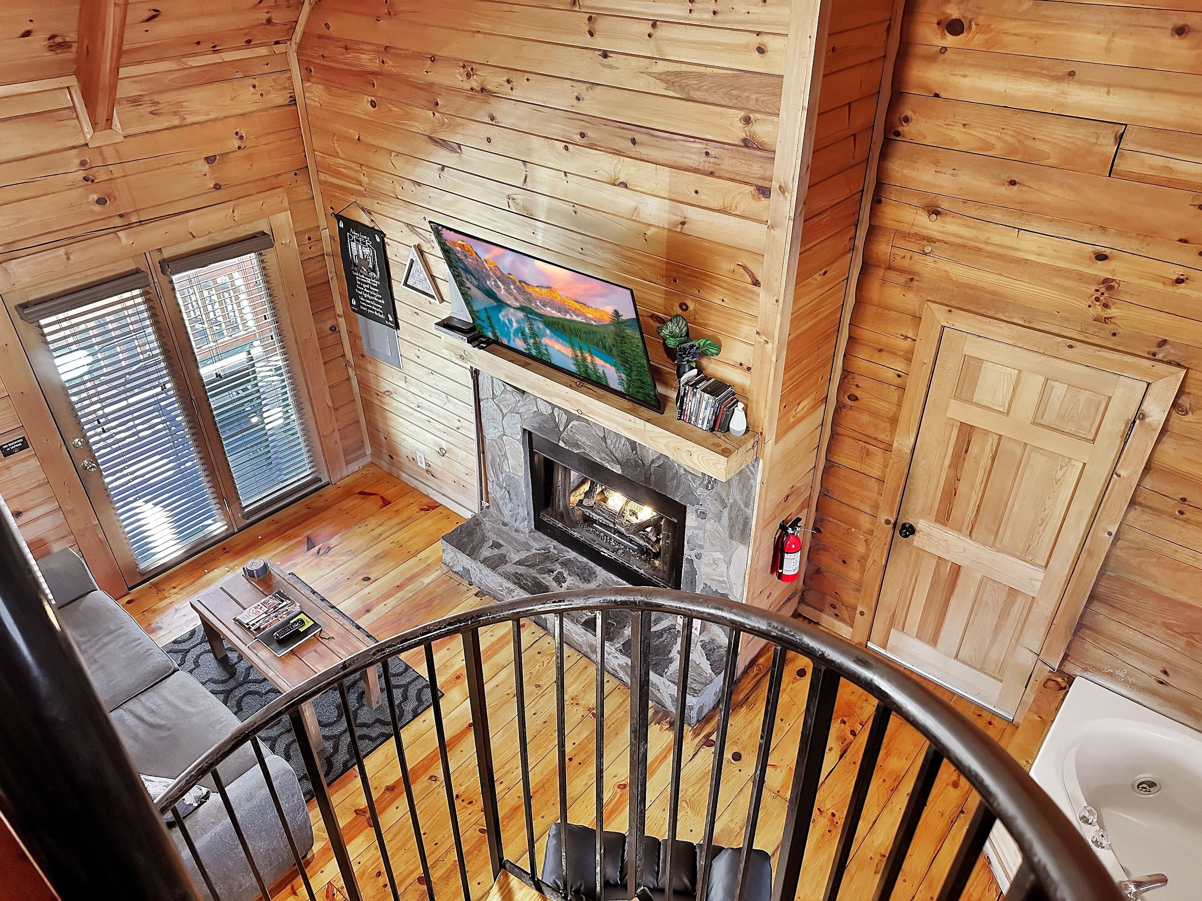 A wooden cabin interior with a stone fireplace, a television mounted above it, and a staircase leading to a balcony.
