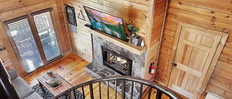 A wooden cabin interior with a stone fireplace, a television mounted above it, and a staircase leading to a balcony.