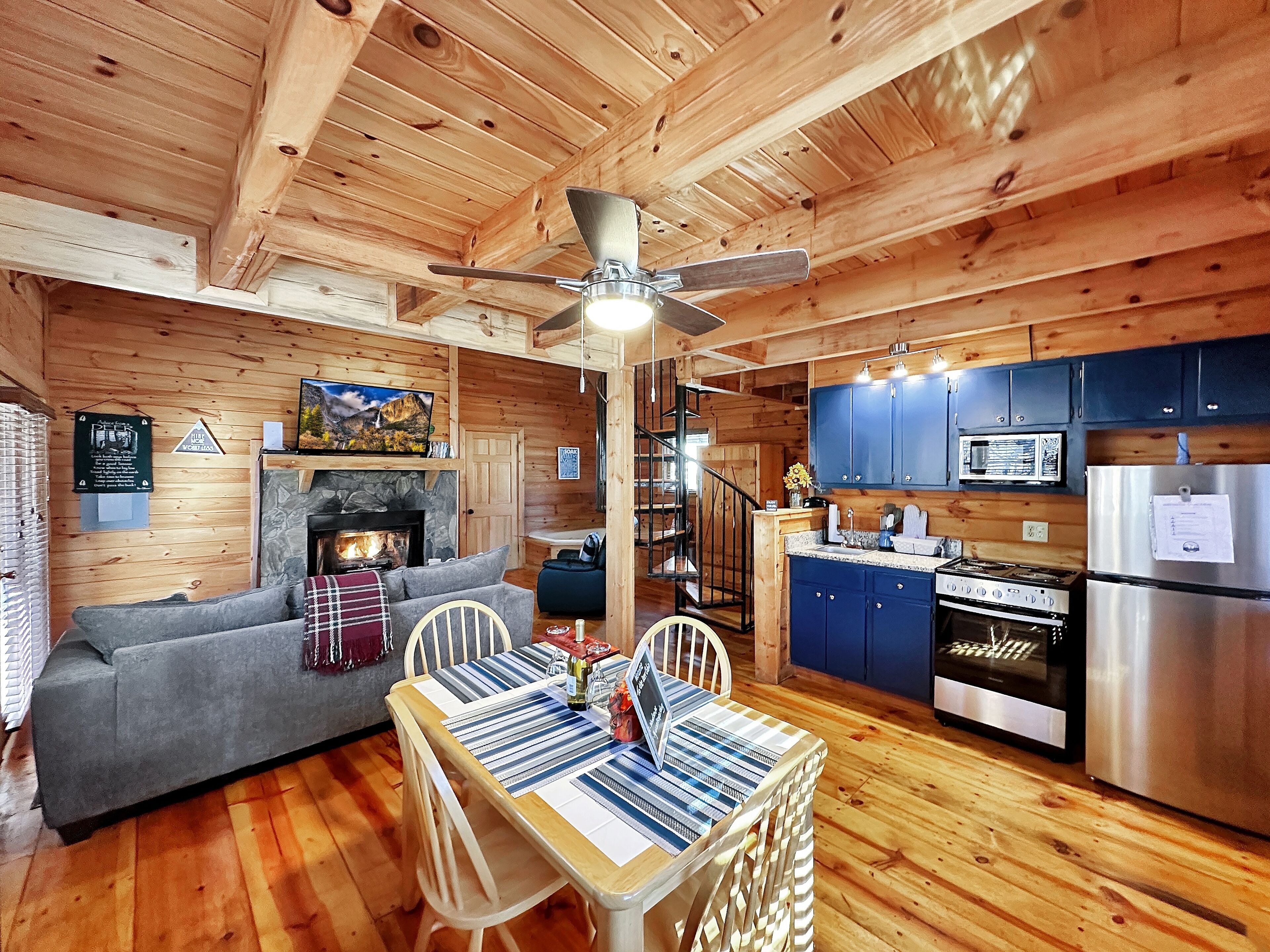 Een gezellig interieur van een blokhut met een open haard, houten wanden, een eethoek en een keuken met blauwe kastjes en roestvrijstalen apparatuur.