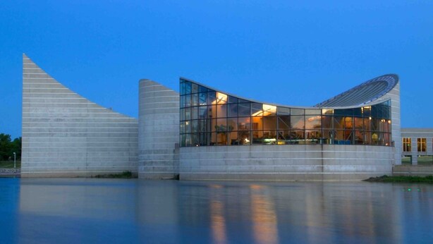 A modern building with a curved glass facade and a reflective water surface in front.