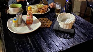 Une personne est assise à une table en plein air et prend son repas : une assiette de nourriture, une bouteille et un verre sont devant elle.