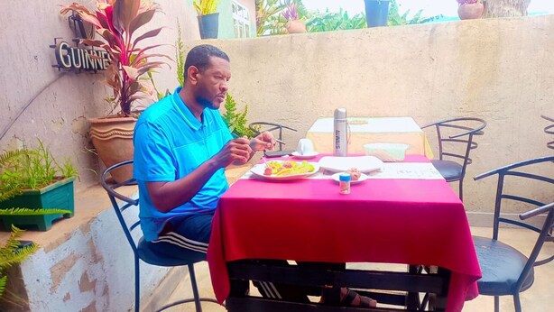 Un homme est assis à une table en plein air, recouverte d’une nappe rouge, et est entouré de plantes en pot. Un panneau indique « GUINNESS ».