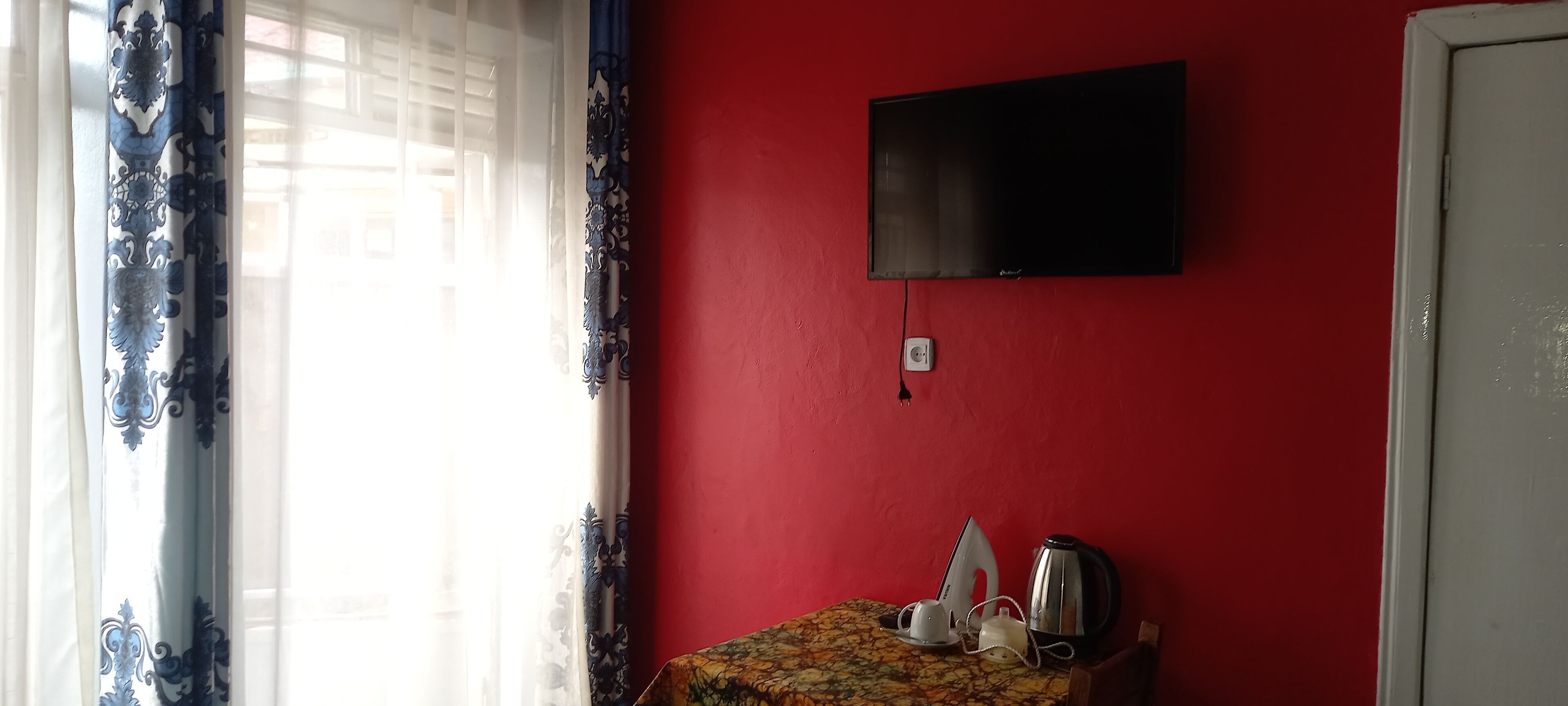 A room with a red wall, a flat-screen TV mounted above a table with a kettle and a cup.