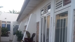 Un bâtiment aux murs extérieurs blancs, doté de nombreuses fenêtres et d’une porte avec un panneau de verre. Des plantes en pot sont disposées le long du mur.