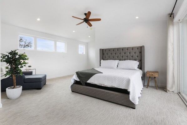 A spacious bedroom with a large bed, a ceiling fan, a potted plant, and a view of the outdoors.