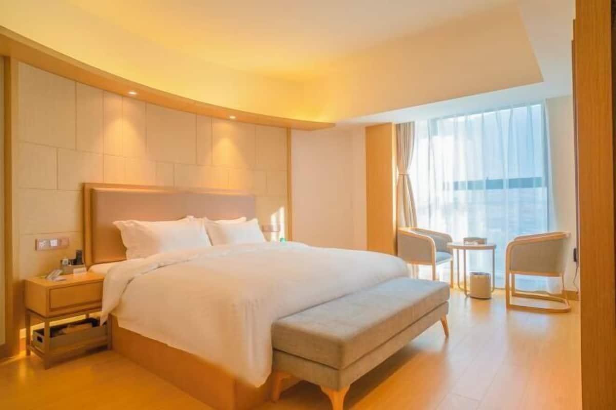 Una habitación de hotel con una cama grande, mesitas de noche, un rincón de descanso con sillas y un ventanal amplio.