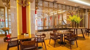 Een restaurant met houten tafels en stoelen, een decoratieve muur met ingewikkelde patronen, en een vaas met gele bloemen.