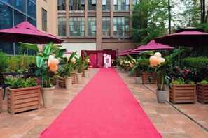 Un tapis rouge menant à l’entrée d’un bâtiment, agrémenté de plantes en pot et de parapluies.