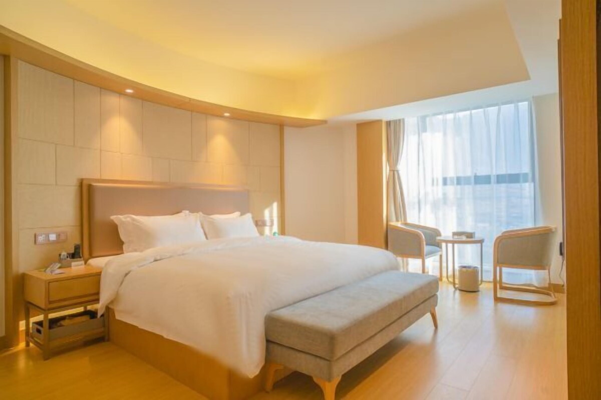 Une chambre d’hôtel moderne dotée d’un grand lit, de tables de chevet, d’un coin salon avec un fauteuil et une petite table, ainsi que d’une grande fenêtre avec des rideaux.