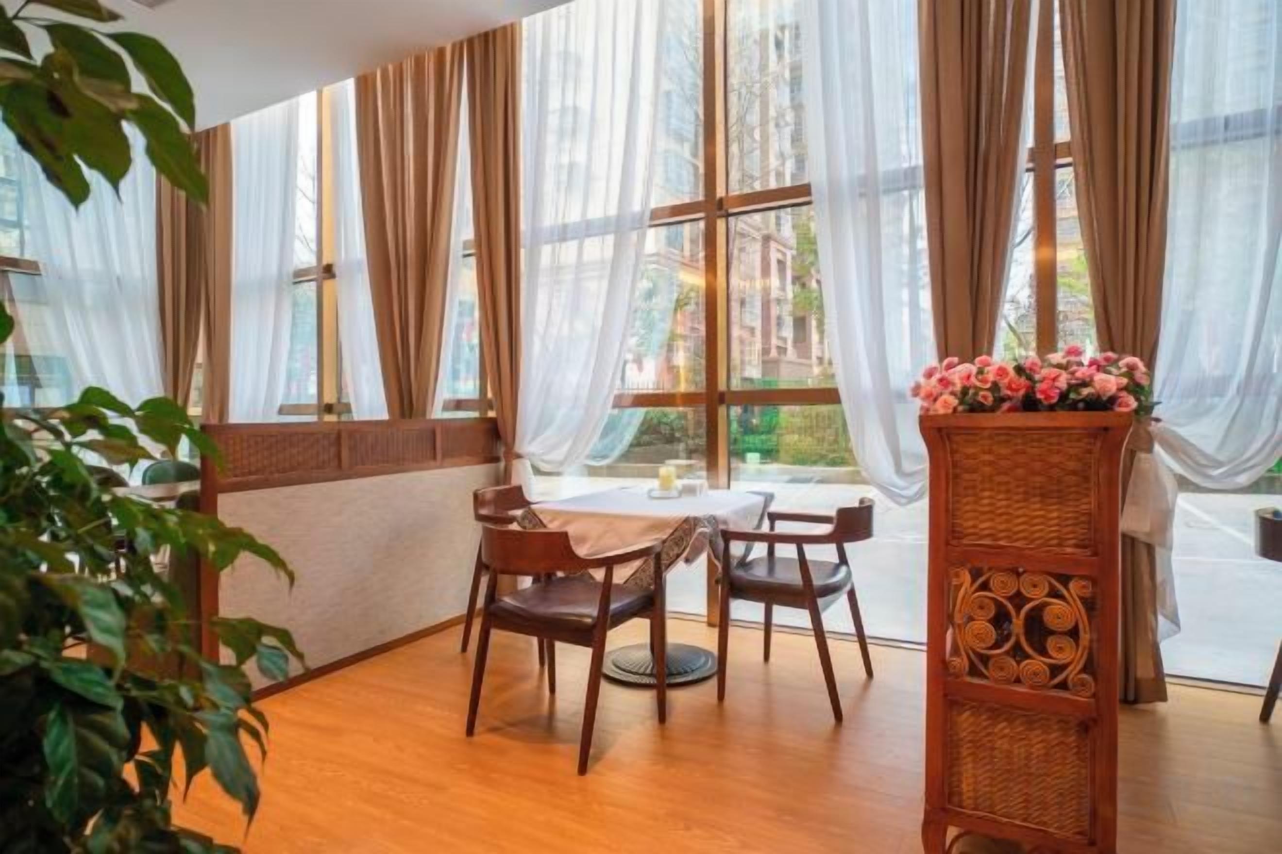 Una habitación con una mesa y sillas, ventanas amplias con cortinas translúcidas y una mesita de mimbre con flores.