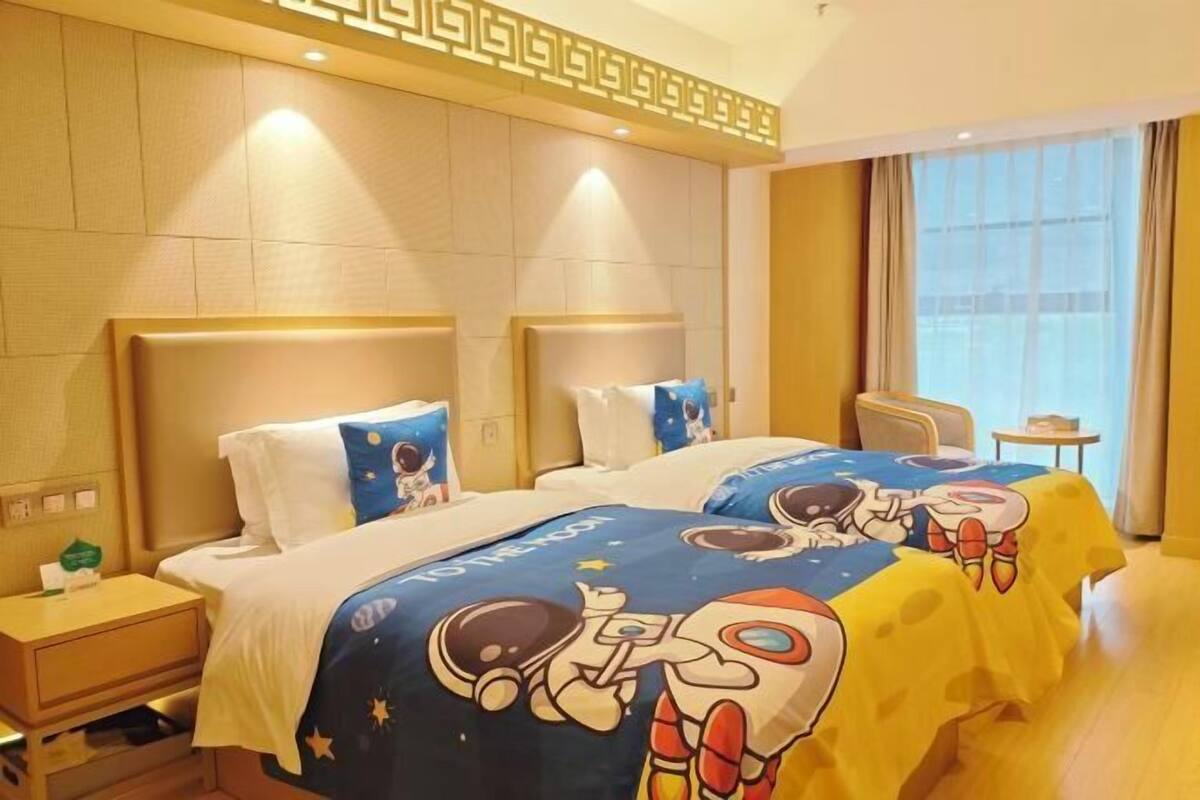 Habitación de hotel con dos camas, cada una con cubrecama con motivos espaciales, una mesita de noche pequeña y un ventanal con cortinas.