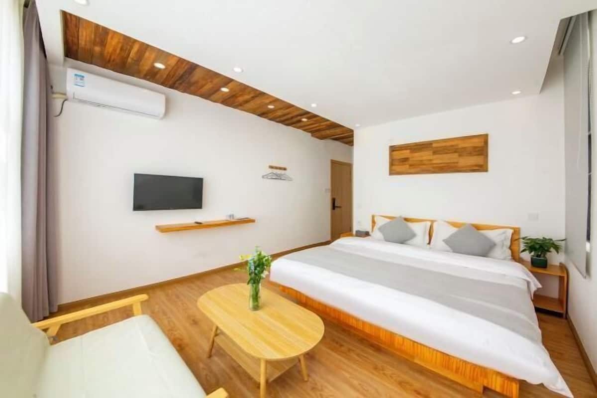 A modern hotel room with a wooden floor, a bed with white bedding, a small round table with a plant, a mounted TV, and a wooden ceiling.
