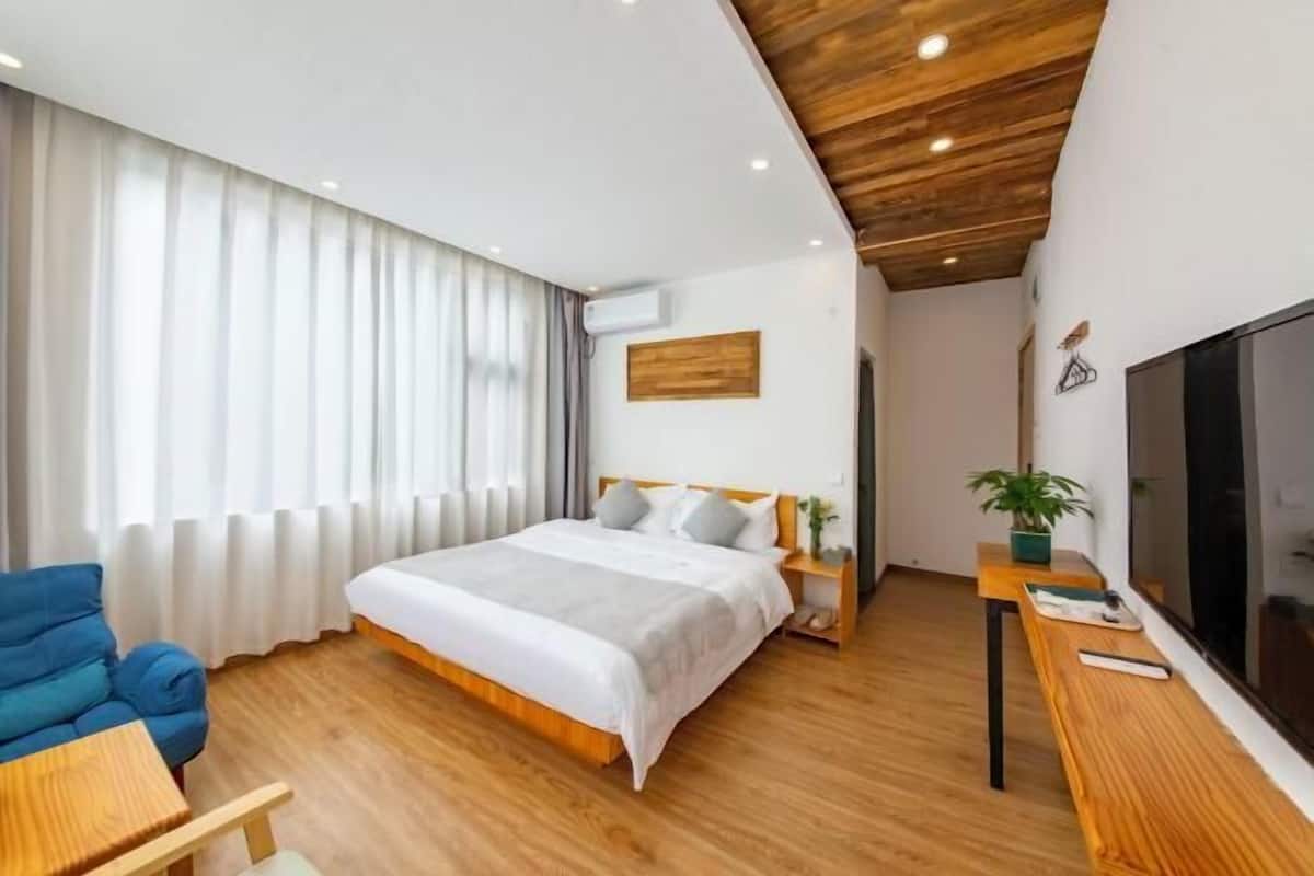 A modern hotel room with a wooden ceiling, a large bed with white bedding, a blue sofa, a wooden side table, and a TV on the right wall.