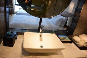 A hotel bathroom with a large sink, a mirror, and a view of a bedroom with a bed and pillows.