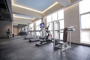 A gym with treadmills, elliptical machines, and weight machines, large windows, and a flat-screen TV.