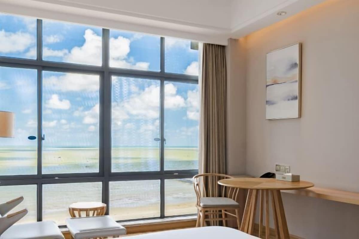 A hotel room with a large window offering a view of the beach and ocean. There is a bed, a small round table with chairs, and a wall-mounted artwork.