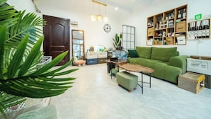 Ein modernes Wohnzimmer mit einem grünen Sofa, einem Holztisch und einem Essbereich. Ein Bücherregal ist mit Büchern und Dekoration gefüllt. Im Vordergrund steht eine große Pflanze.