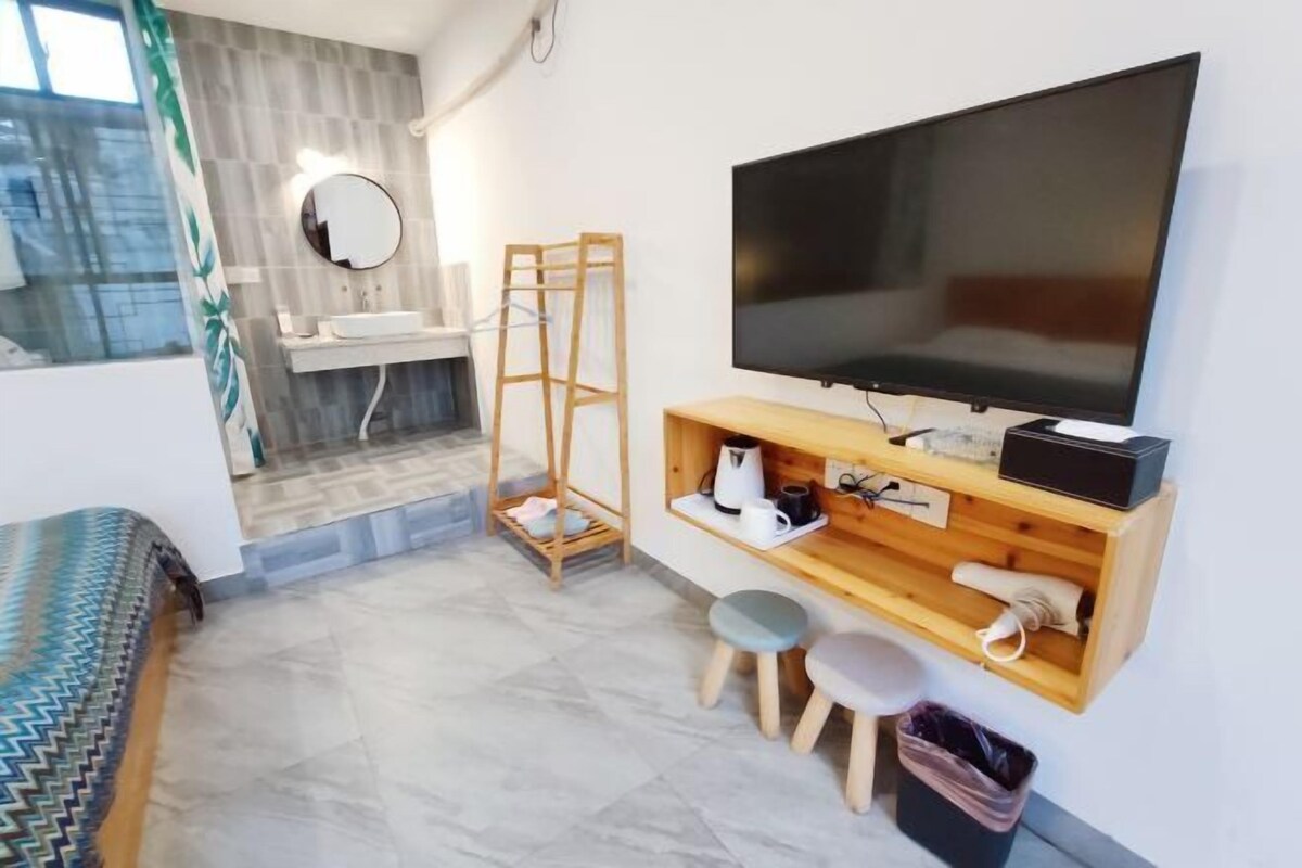 A compact room with a bed, a TV, a wooden shelf with items, a mirror, and a window with curtains.