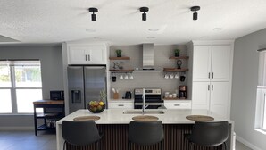 A modern kitchen with white cabinets, a central island with bar stools, stainless steel appliances, and a window providing natural light. - The Ultimate Escape (Palm Coast)