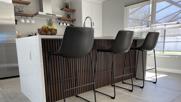 A modern kitchen with a central island, two black bar stools, and a window providing natural light. - The Ultimate Escape (Palm Coast)