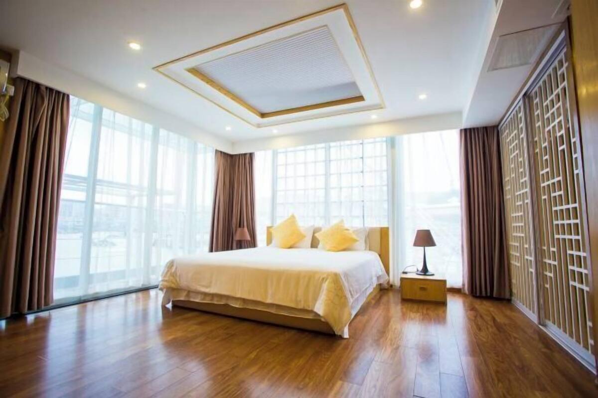 A spacious hotel room with a large bed, a nightstand, and a view through floor-to-ceiling windows.