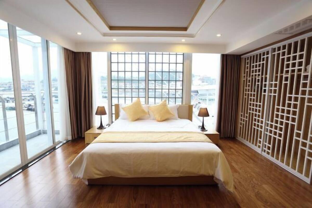A spacious hotel room with a large bed, two bedside tables, and a view of the city through floor-to-ceiling windows.