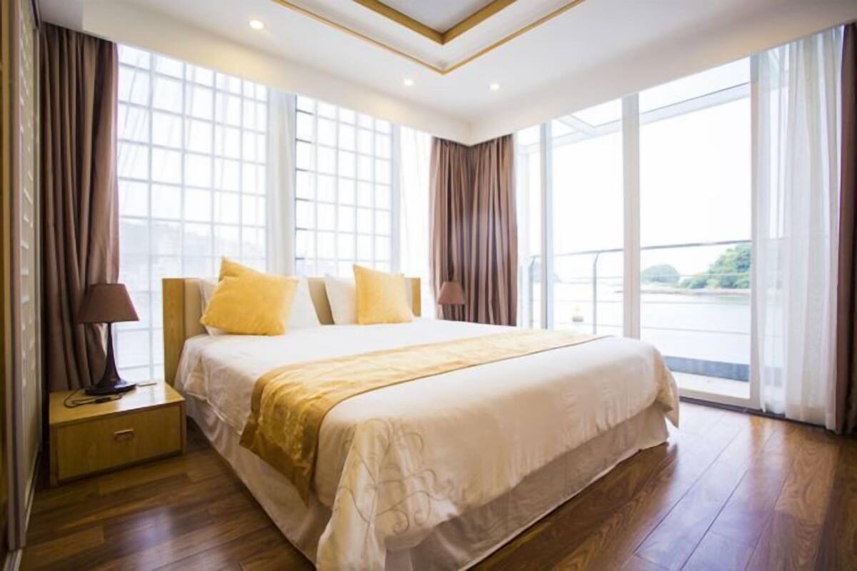 A spacious bedroom with a large bed, a nightstand, and a view of a body of water through large windows.
