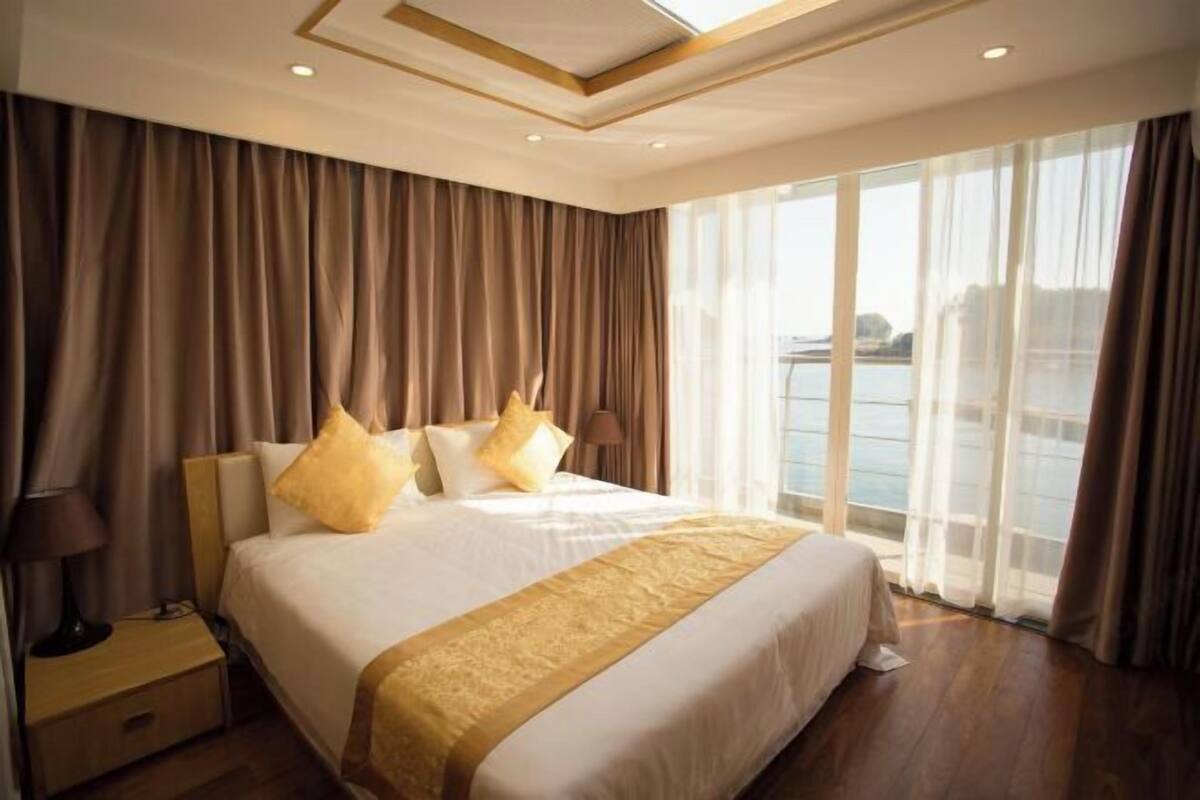 A hotel room with a large bed, a nightstand, and a view of the sea through large windows.