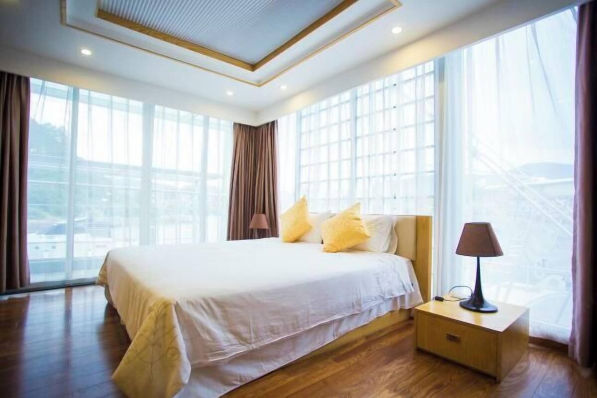 A hotel room with a large bed, a bedside table with a lamp, and large windows with curtains.