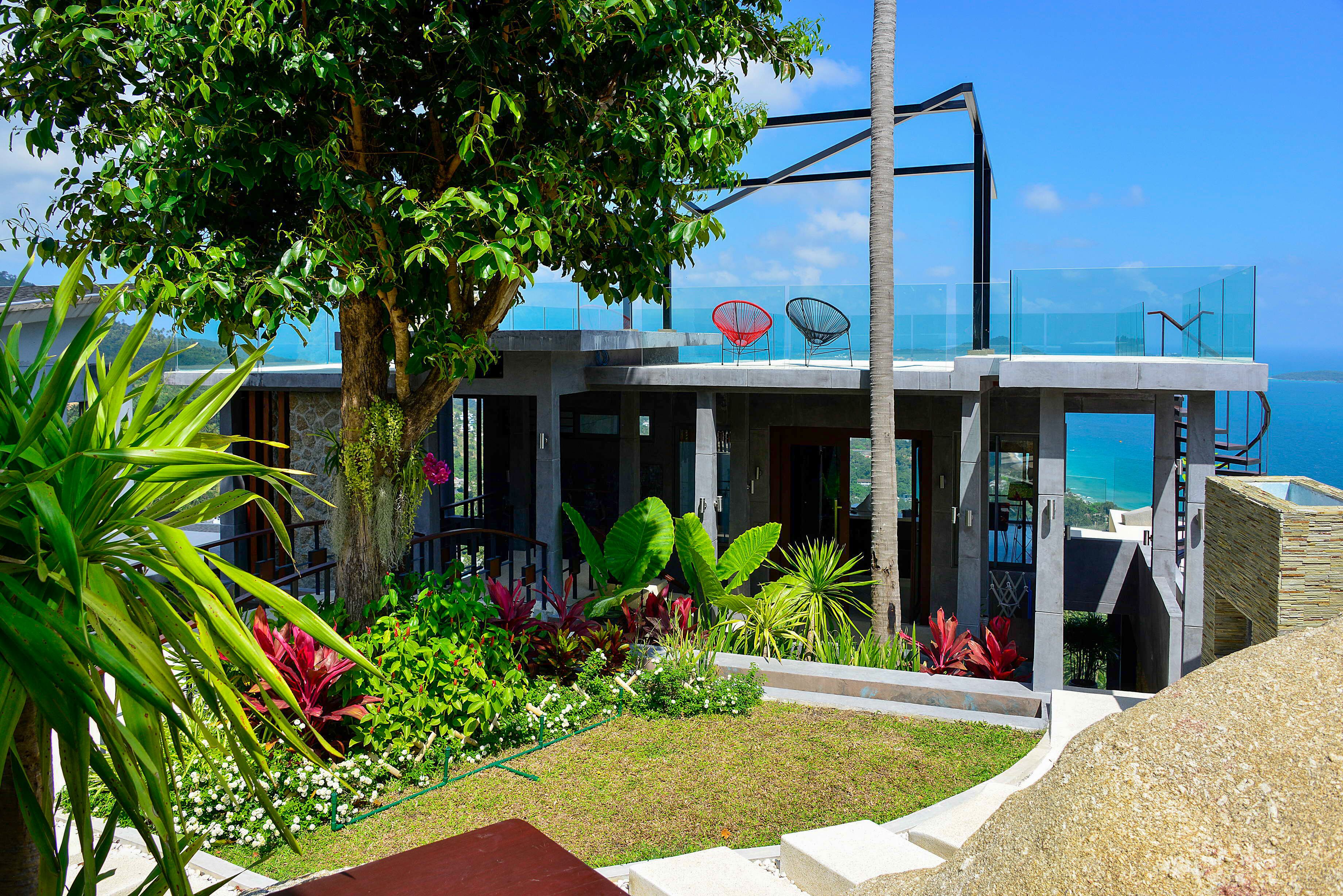 A modern building with a glass balcony overlooking the sea, surrounded by lush tropical plants.