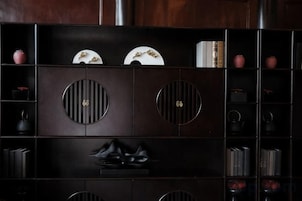 Um armário de madeira escura com recortes circulares, prateleiras com livros e objetos decorativos, e um par de sapatos pretos na prateleira inferior.