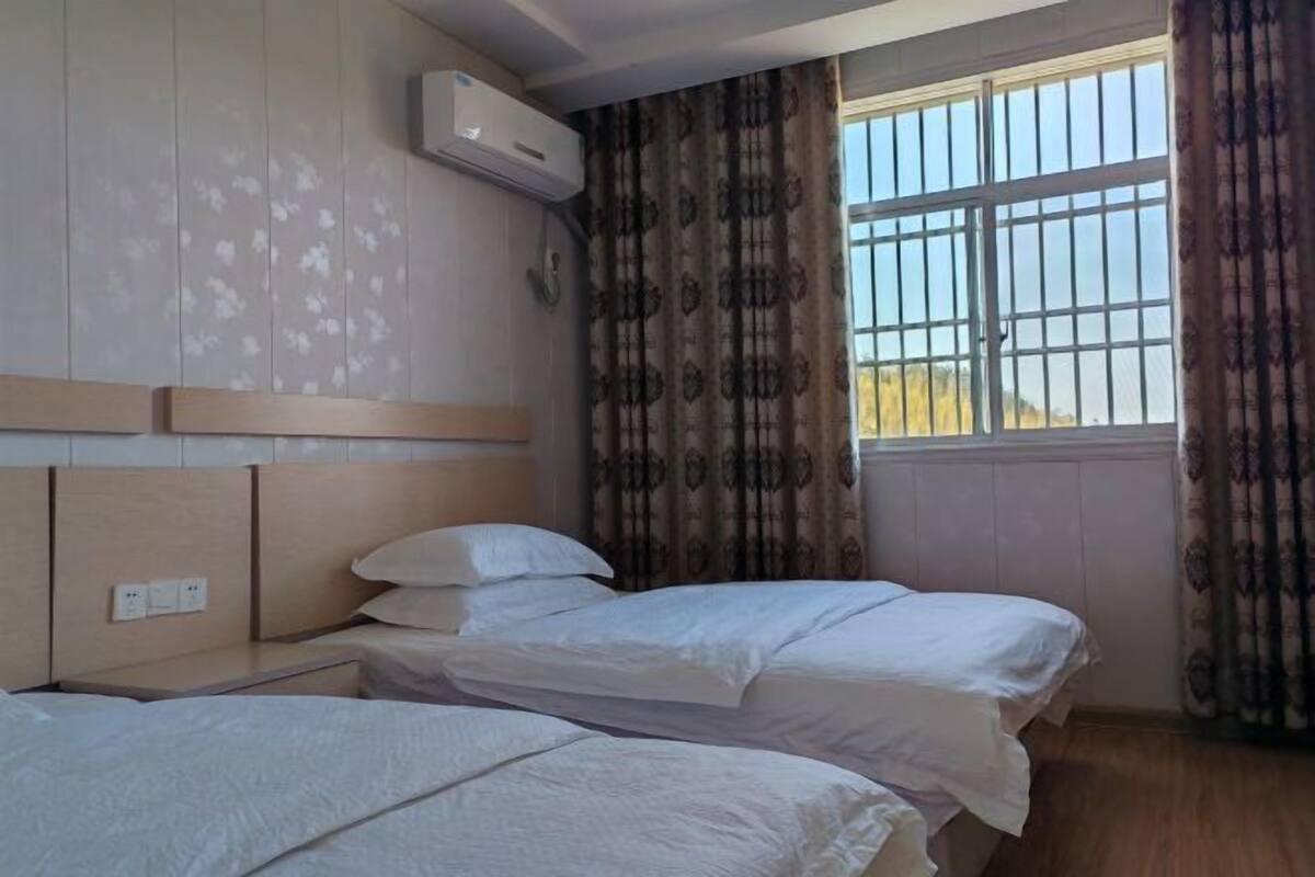 A hotel room with two beds, a window with bars, a patterned curtain, and an air conditioning unit on the wall.
