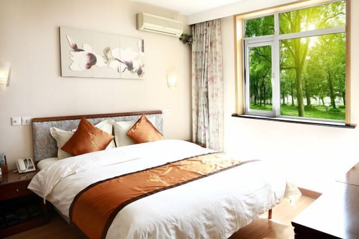 A bedroom with a bed, a nightstand, a telephone, a window with a view of trees, and a wall-mounted air conditioner.
