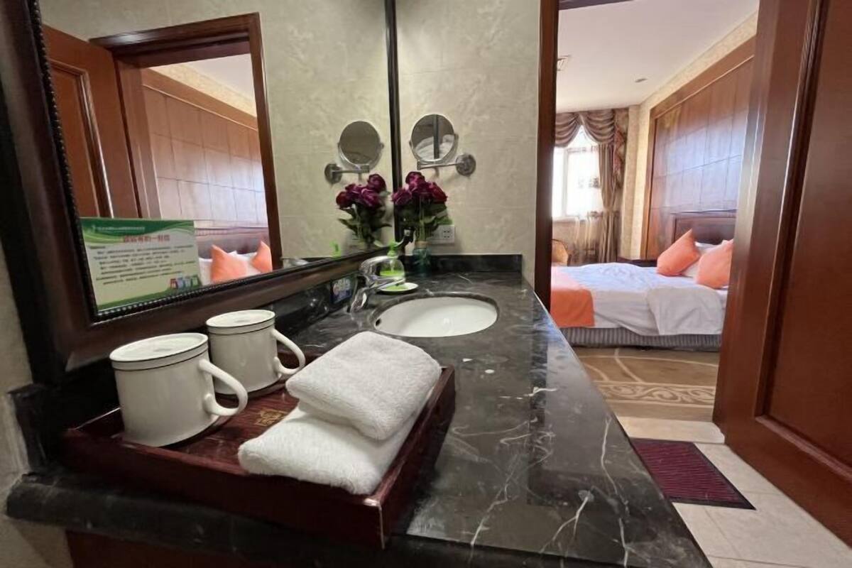 Une salle de bain d’hôtel avec un comptoir en marbre, deux lavabos, un miroir et un lit visible à travers une porte ouverte.