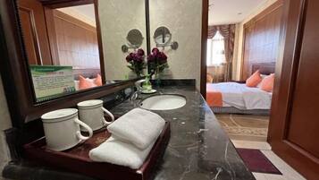 Ein Hotelbad mit Marmorplatte, zwei Waschbecken, einem Spiegel und einem durch eine geöffnete Tür erkennbaren Bett.