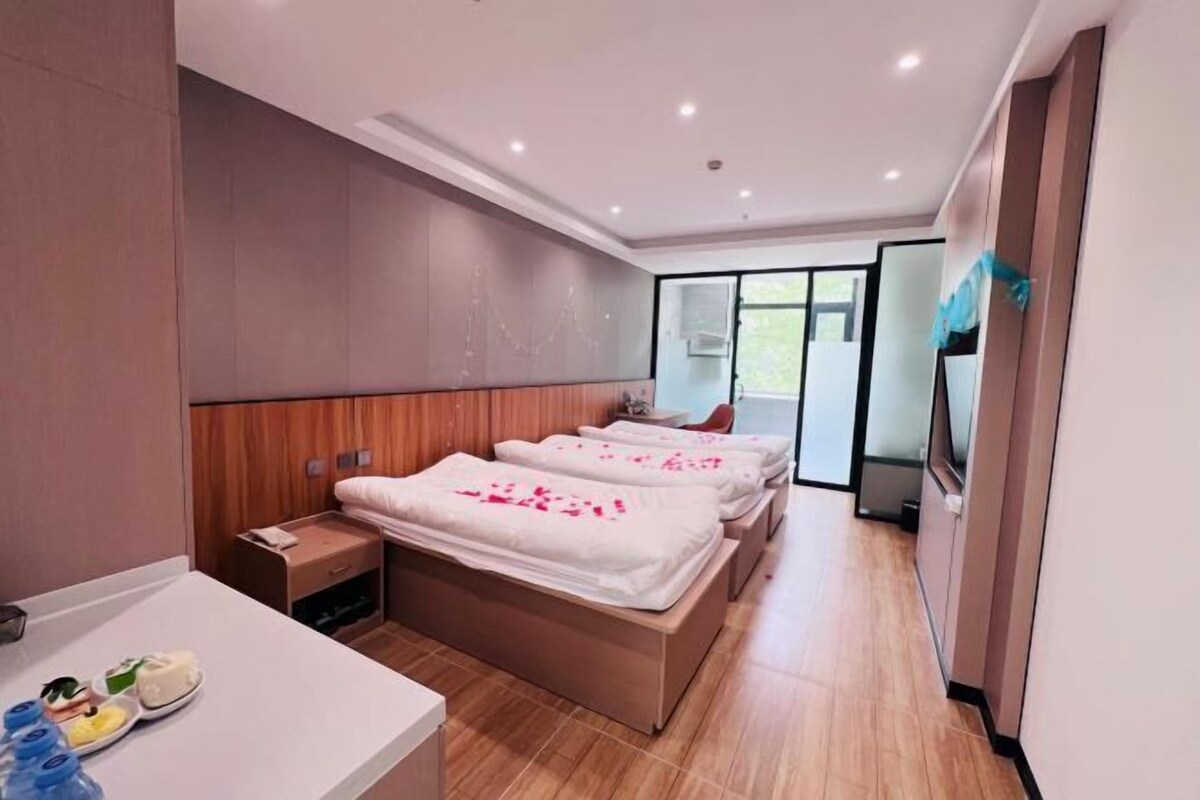 A modern hotel room with a wooden headboard, a large bed with white bedding, a bedside table with a lamp, and a view of the outdoors through a glass door.