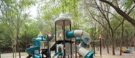 A playground with slides and a climbing structure surrounded by trees.
