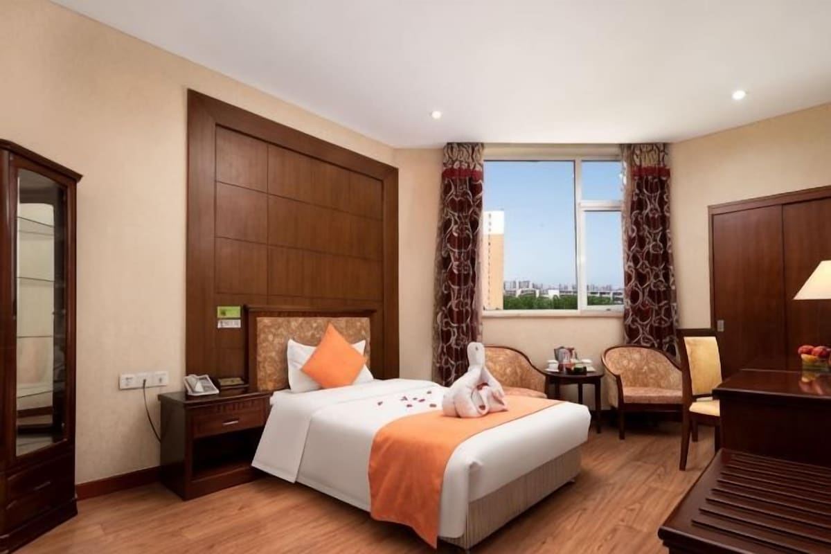 Een hotelkamer met een groot bed, een bureau, een stoel, een raam met gordijnen en uitzicht op de stad.