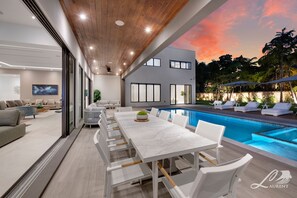 A modern outdoor dining area with a pool, white furniture, and a sunset view.