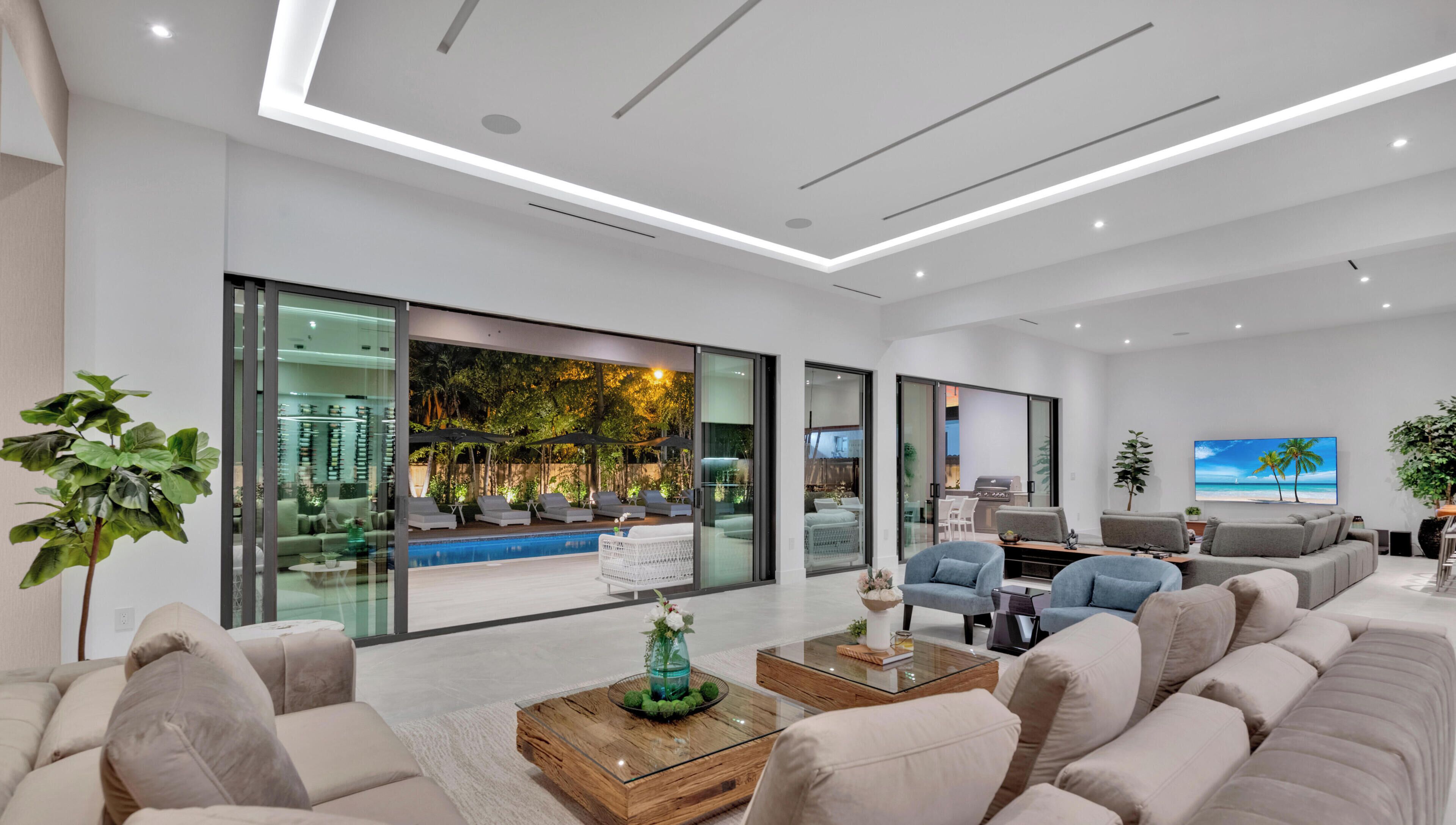 Sala de estar moderna com um sofá grande, uma mesa de centro e uma televisão. Há uma parede de vidro com vista para uma piscina e móveis de exterior.
