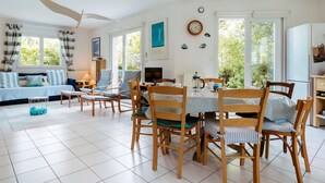 A spacious living area with a dining table, chairs, and a sofa. There are large windows with curtains, a ceiling fan, and a clock on the wall. - Carnac - 4-room house - 102m² - Wifi - 2* (CARNAC)