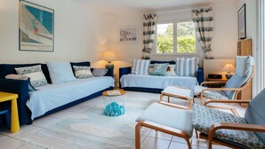 A cozy room with a blue sofa, a wooden chair, a small table, and a window with curtains. - Carnac - 4-room house - 102m² - Wifi - 2* (CARNAC)