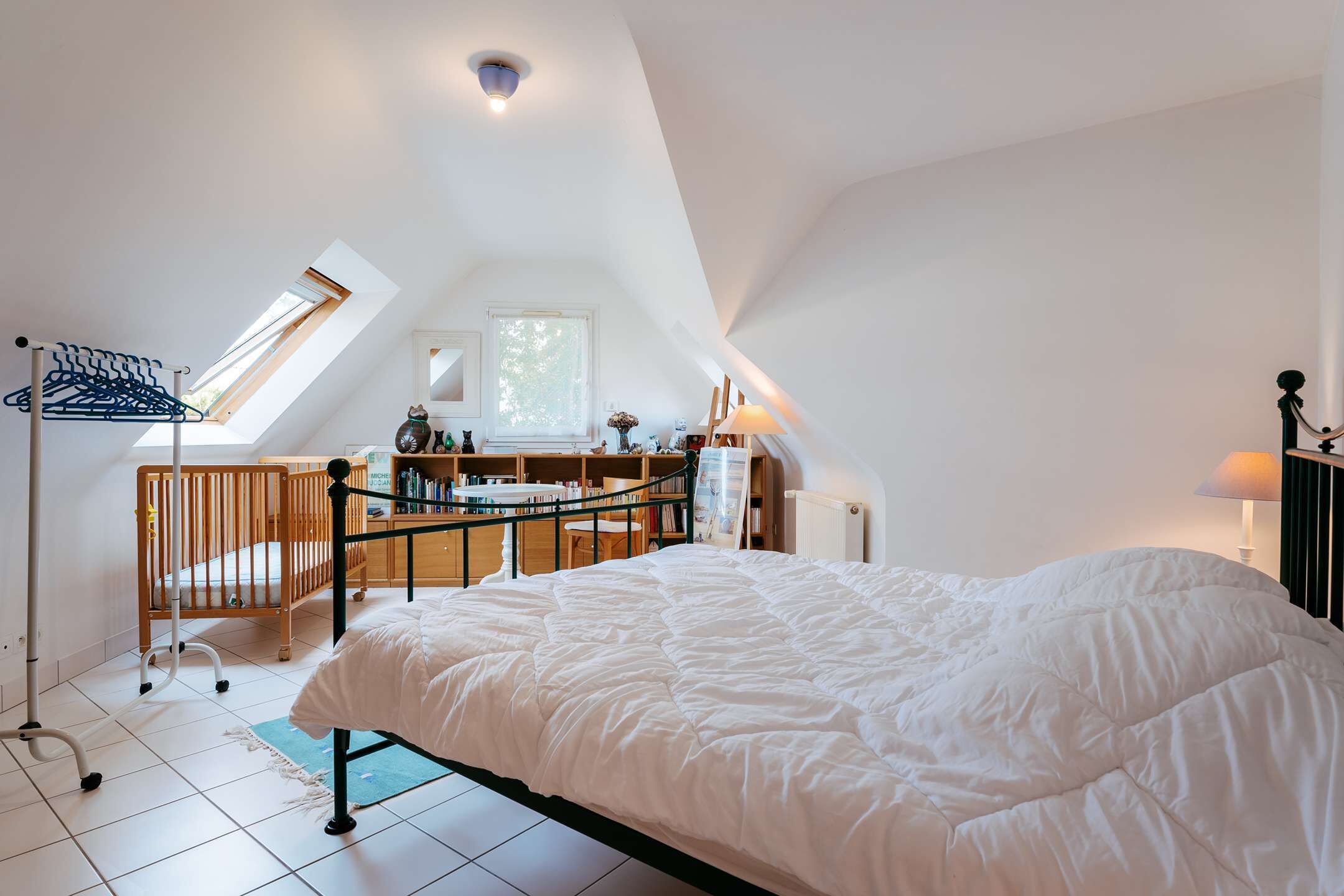 A bedroom with a large bed, a crib, a desk with a chair, and a window with a view of trees.