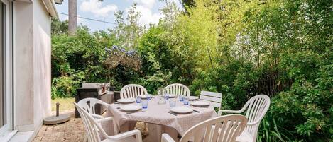 Une terrasse extérieure avec une table dressée pour quatre personnes, entourée de chaises en plastique blanches, le tout se fondant dans un décor de verdure luxuriante et d’une maison.
