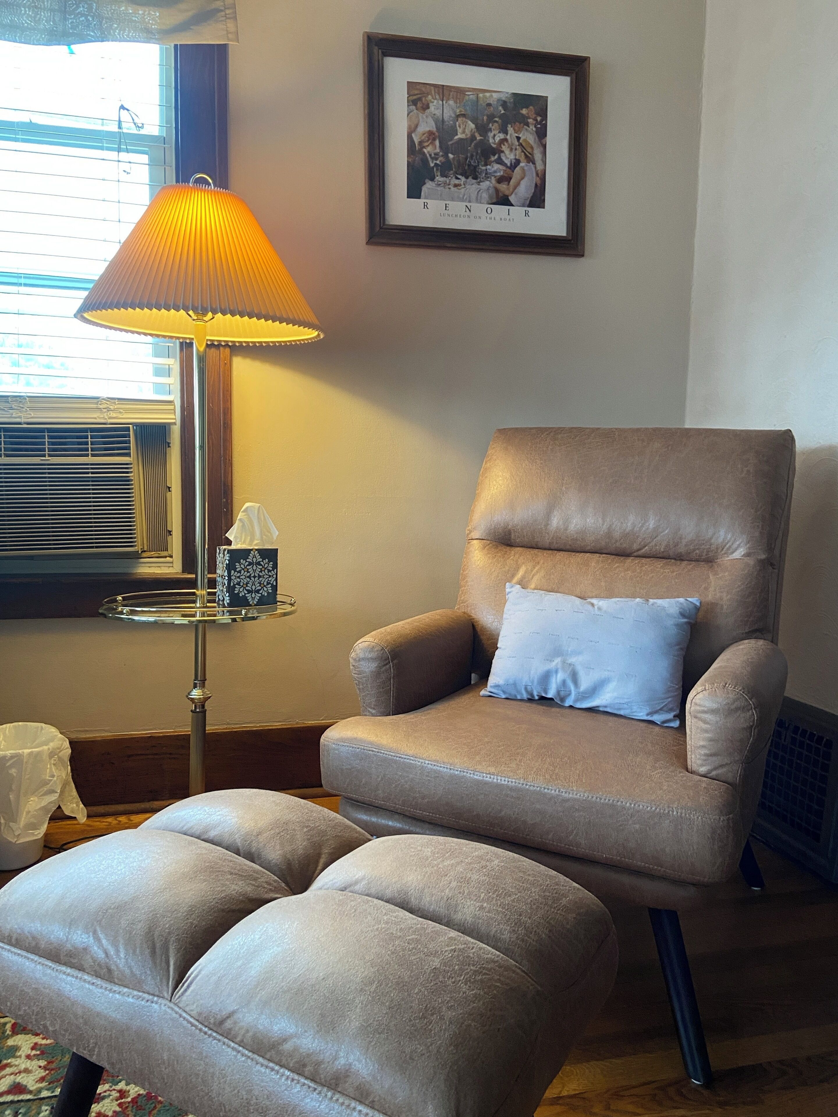 A living room with a brown leather armchair, a matching ottoman, a floor lamp, a framed picture on the wall, and a window with blinds.