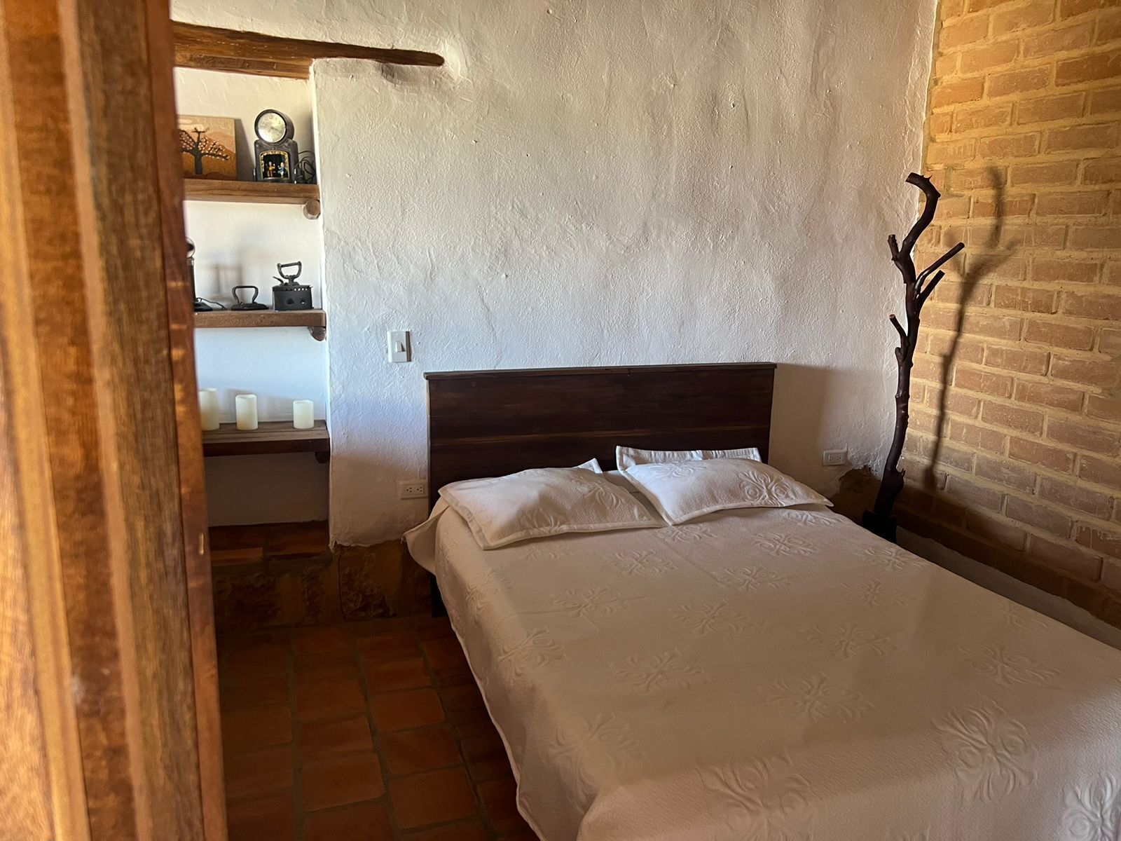 Een slaapkamer met een houten hoofdbord, een bed met witte lakens, een bakstenen muur en een plank met diverse spullen.