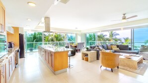 A modern kitchen with a central island, a dining area with a sofa and chairs, and large windows offering a view of palm trees and the ocean. - Península T3 1A Luxurious Ocean View Getaway! (Puerto Vallarta)