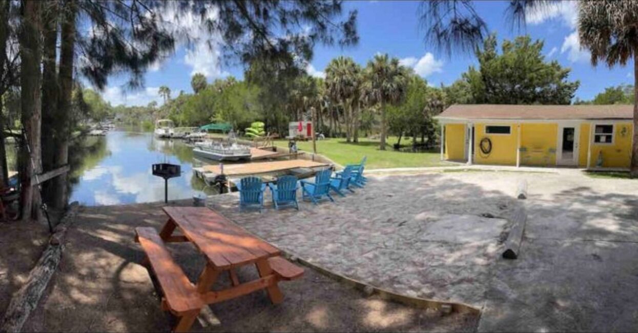 A sandy area with a picnic table and blue chairs, a small yellow building, and a calm water body with boats.